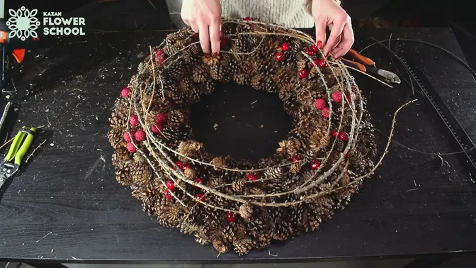 A finished pinecone wreath on a worktable, with small red decorations and slender larch twigs following the circle.