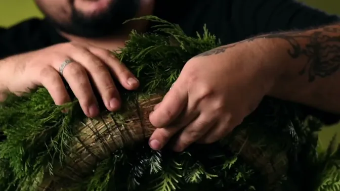 Hands beside a wire hanging loop on the exposed hay back of a mixed-greenery wreath