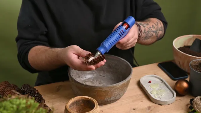Hot glue being applied along the cut face of a spruce-cone half beside an empty bowl.
