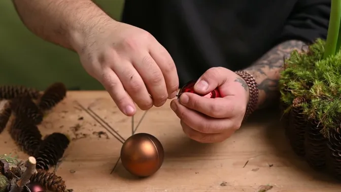 Hands wrapping stiff wire around the neck of a red plastic bauble, with a bronze bauble on a wire stem nearby.