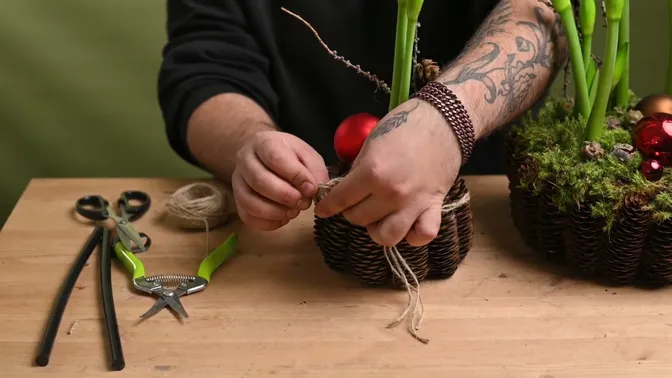 Hands tying several strands of natural twine into a bow around the smaller cone-covered planter.