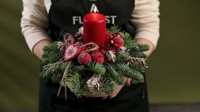 Red candle surrounded by evergreen branches, red baubles, berries, cones and a jute bow.