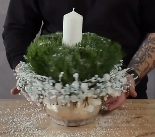 Round pine arrangement with a white candle, pale cones and white decorations in a pot.