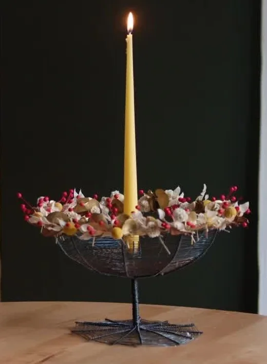 Woven wire bowl on a raised stem, filled with dried flowers and red accents around a slender yellow candle.