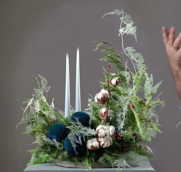 Asymmetrical evergreen arrangement with two white candles, blue baubles, cotton and cones in a low dish.