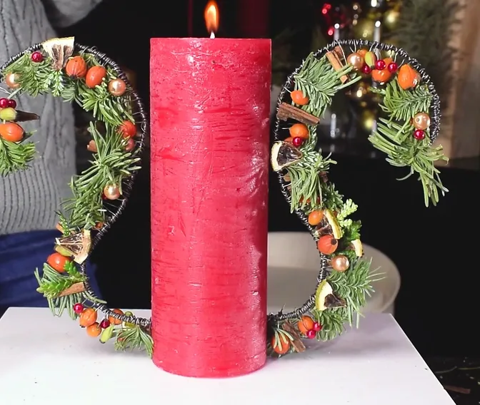 Red pillar candle between two curved evergreen wings decorated with orange and red accents.
