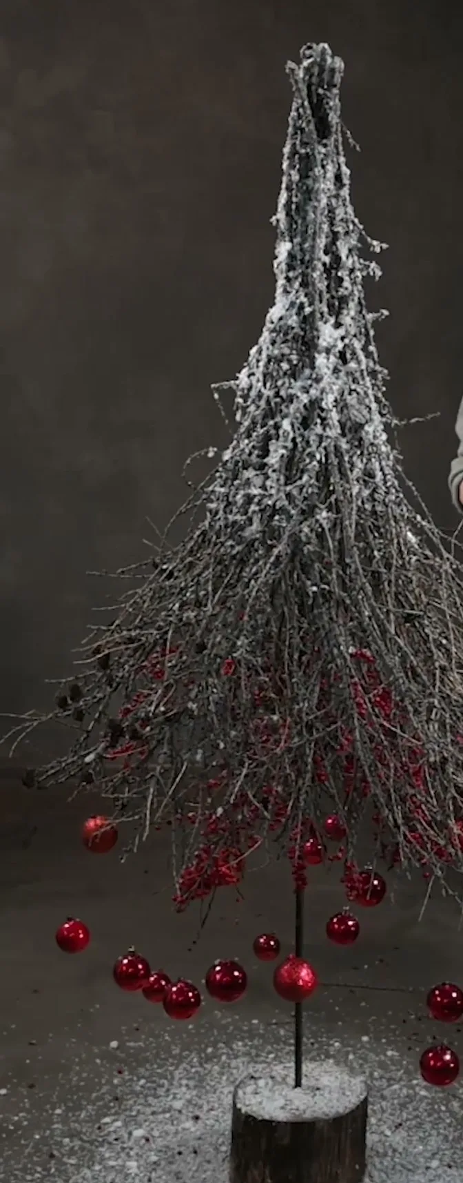 Tall snowy branch Christmas tree with red hanging baubles on a round wood base.