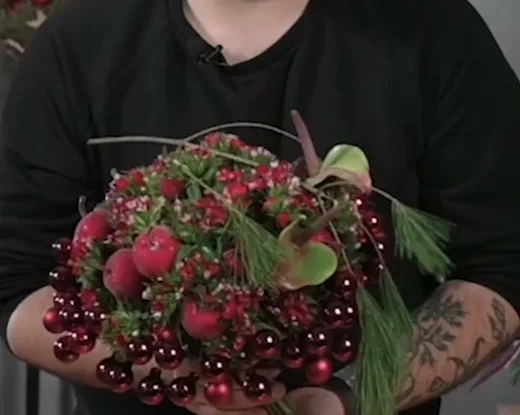 Round Christmas bouquet of red baubles, red flowers and evergreen branches held in two hands.