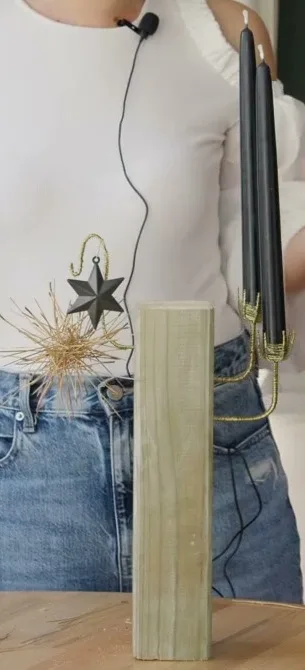 Two black candles in woven wire cups extend from an upright wooden block, with a dry pine-needle ornament and dark star on the other side.